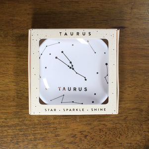 The Taurus ring dish from Lucky Feather on a wood background. The dish is cream colored with the word "TAURUS" and the Taurus star constellation printed on it.