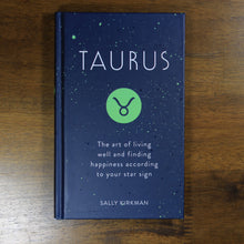 Load image into Gallery viewer, An overhead view of book: "Taurus: The art of living well and finding happiness according to your star sign" by Sally Kirkman, on a wood background.