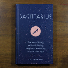 Load image into Gallery viewer, A dark blue book titled Sagittarius: The art of living well and finding happiness according to your star sign. The book is by Sally Kirkman. The cover has the title, author, and the Sagittarius zodiac symbol in a light pink circle.