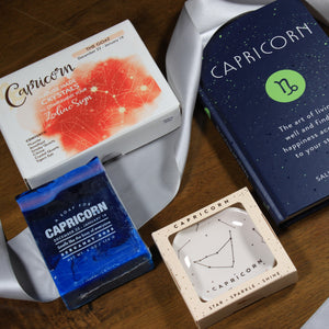 Photo shows the contents of the What's Your Sign Capricorn Gift Box: Capricorn book, Capricorn ring dish, Capricorn crystals, and Capricorn soap. Items are laid over a silver ribbon.