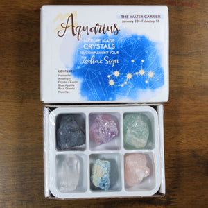 Aquarius Horoscope Crystals from Rock Paradise. Photo shows a white box that has a blue colored ink blot with the Aquarius constellation and illustrations of gemstones. The box reads " Aquarius nature made crystals to complement your zodiac sign. The Water Carrier. January 20-February 18. Contents: Hematite, Amethyst, Crystal Quartz, Blue Apatite, Rose Quartz, Fluorite."