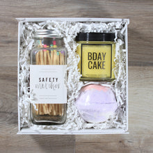 Load image into Gallery viewer, The Mini Birthday gift box from Doromania: a glass jar of matches with multicolor tips, a yellow candle with a black lid that says "Bday Cake", anda purple bath bomb all in a white box with white crinkle paper