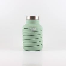 Load image into Gallery viewer, A photo of the compressed version of an expandable dusty sage water bottle with a metal cap, that says "que" in white lowercase letters.