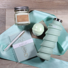 Load image into Gallery viewer, A flat lay of the contents of the "For Your Enjoy-Mint" gift box, which includes a mason jar candle with light green wax, a light green bath bomb, a light green water bottle with a metal cap, a light green journal that says "bucket list", and a white pen with gold accents. The items are laid across a mint green ribbon on a wood floor.