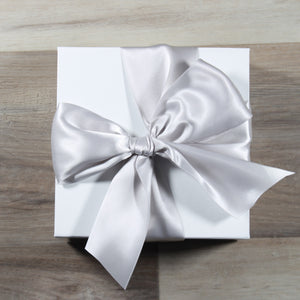 An overhead photo of a Doromania gift box - a white square gift box with a silver satin bow on a wood background.
