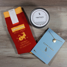 Load image into Gallery viewer, Flat lay photo of the contents of the A Little Bit Capricorn gift box: Capricorn candle, Capricorn socks, and a Capricorn Necklace. A fun horoscope gift for friends.