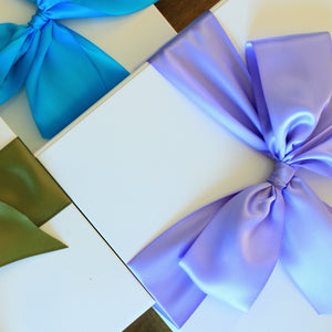This photo shows an overhead view of three white gift boxes, each with different colored satin ribbon tied in a bow. The bows are purple, blue, and green.