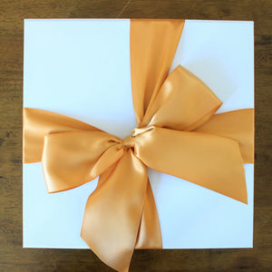 This photo is an overhead view of white gift box with butterscotch orange satin ribbon tied in a bow, on top of a wood table