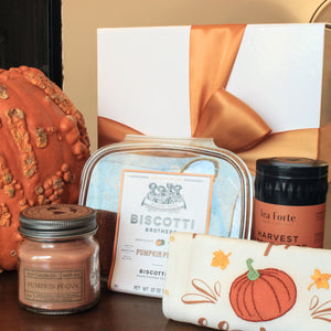 In this photo is a group of items from the "Fall Fun" seasonal gift box, including a brown mason jar candle, a plastic container of biscotti, a tea towel with a pumpkin, and a tin of loose tea. They are placed in front of a pumpkin and a white gift box with dark orange ribbon.