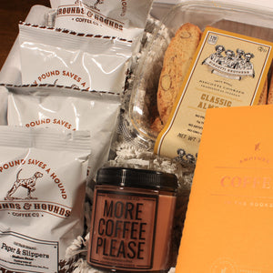 This photo is a close up of the contents of the Doromania "Coffee on Coffee" gift box, which includes a brown coffee candle, a yellow coffee journal, a plastic container of biscotti, and four white bags of coffee grounds.