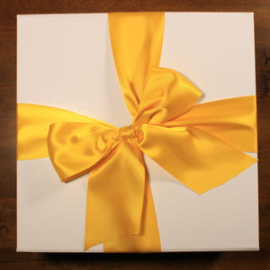 This photo shows an overhead view of a white gift box with yellow satin ribbon tied in a bow, on top of a wood table
