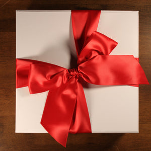 This photo shows an overhead view of white gift box with red satin ribbon tied in a bow, on top of a wood table
