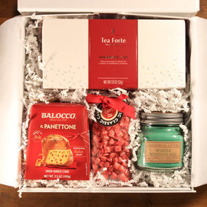 This photo shows the Doromania "Home for the Holidays" gift box, which includes a green candle, a red and white box of winter tea, a mini panettone, and peppermint all in a white box with white crinkle paper.