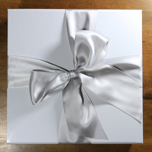 An overhead view of a white magnetic square gift box with a silver satin ribbon tied in a bow