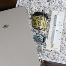 Load image into Gallery viewer, This photo is an overhead shot of a white gift box filled with white crinkle paper that has a white pen, a light blue essential oils container, a yellow candle and a grey planner notebook. The planner is being held over the top of the box and the other three items are inside the box.