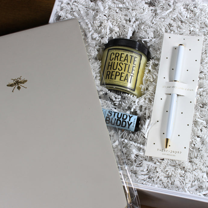 This photo is an overhead shot of a white gift box filled with white crinkle paper that has a white pen, a light blue essential oils container, a yellow candle and a grey planner notebook. The planner is being held over the top of the box and the other three items are inside the box.