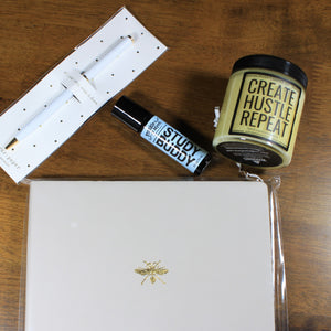 This is an overhead shot of the contents of the "Every Day I'm Hustlin'" office gift box: a white twist pen, a light blue peppermint aromatherapy roller, a yellow candle, and a grey journal planner.
