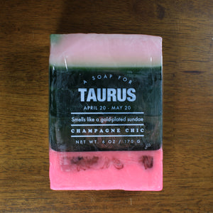 A Soap for Taurus from Whiskey River Soap Co. on a wood background. The soap is colored in thirds with pink on the bottom, green in the middle, and white on the top.