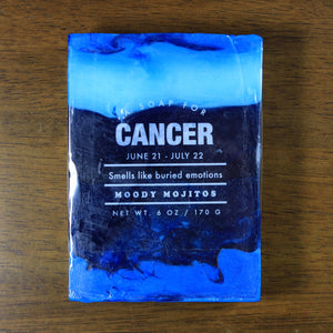 Whiskey Rivers Soap Co. Cancer Astrology Soap. The bar is three colors of blue swirled together. In white writing, says "a soap for Cancer. June 21-July 22. Smells like buried emotions. Moody Mojitos."