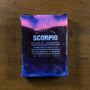 Whiskey Rivers Soap Co. Scorpio Astrology Soap. The bar is dark blue with pink marbled into the top. In white writing, says "a soap for scorpio. October 23-November 21. Smells like a Supervillain. Vindictive Violets."
