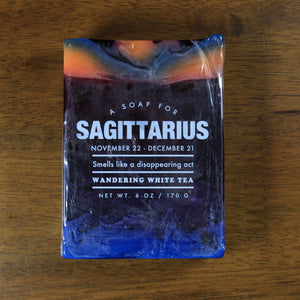 Whiskey Rivers Soap Co. Sagittarius Astrology Soap. The bar is dark purple with orange and blue marbled into the top. In white writing, says "a soap for Sagittarius. November 22-December 21. Smells like a disappearing act. Wandering White Tea."."