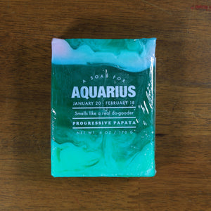 Whiskey Rivers Soap Co. Aquarius Astrology Soap. The bar is a mix of translucent and opaque teal colors. In white writing, says "a soap for Aquarius. January 20-February 18. Smells like a real do-gooder. Progressive Papaya."