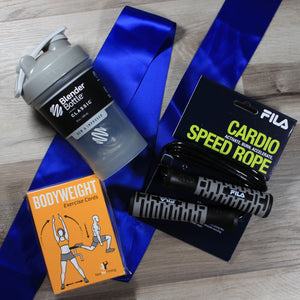 This photo shows a flat lay view of the contents of the "Reps and Ropes" gift box on a wood floor - a grey blender bottle, a black and grey cardio speed rope, and a yellow box of bodyweight exercise cards laying on top of a royal blue ribbon.
