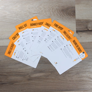 This photo shows eight bodyweight fitness cards spread out in a fan across a wood floor. Each card has a yellow band across the top with the name of an exercise, and the rest of the card is white with written exercise instructions.