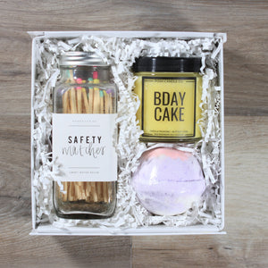 The Mini Birthday gift box from Doromania: a glass jar of matches with multicolor tips, a yellow candle with a black lid that says "Bday Cake", anda purple bath bomb all in a white box with white crinkle paper