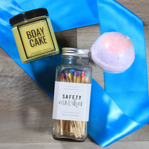 The contents of the MIni Birthday gift box from Doromania: a yellow candle with a black lid that says "bday cake", a purple bath bomb, and a glass jar of matches with multi colored tips, all on top of a teal ribbon on a wood floor.