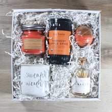 Load image into Gallery viewer, An overhead view of the &quot;Sweater Weather&quot; gift box, which includes a mug that says sweater weather, a bottle of white tipped matches, a red and brown bath bomb, a can of harvest apple spice tea, and a red mason jar candle, all in a white box with white crinkle paper