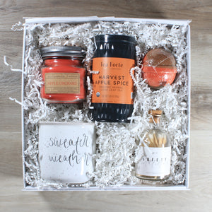 An overhead view of the "Sweater Weather" gift box, which includes a mug that says sweater weather, a bottle of white tipped matches, a red and brown bath bomb, a can of harvest apple spice tea, and a red mason jar candle, all in a white box with white crinkle paper
