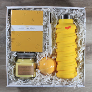 An overhead shot of the "You're The Zest" yellow themed gift box, which includes a yellow journal, a white pen, a yellow water bottle, a yellow bath bomb, and a yellow candle all in a white box with white crinkle paper