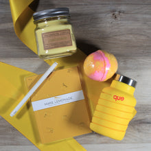Load image into Gallery viewer, An overhead view of the contents of the &quot;You&#39;re the Zest&quot; yellow themed gift box on a wood floor, including a yellow candle, yellow water bottle, yellow and pink bath bomb, yellow journal, and white pen over a yellow ribbon.
