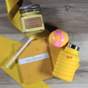 An overhead view of the contents of the "You're the Zest" yellow themed gift box on a wood floor, including a yellow candle, yellow water bottle, yellow and pink bath bomb, yellow journal, and white pen over a yellow ribbon.