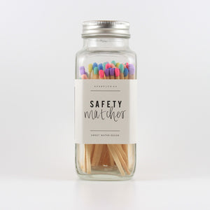 A glass jar with a metal lid, filled with match sticks that have multi colored tips, and a label on the bottle that says "safety matches"