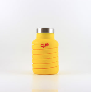 A photo of the compressed version of an expandable citrus yellow water bottle with a metal cap, that says "que" in pink lowercase letters.
