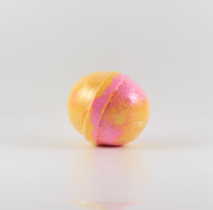 A mostly yellow colored, oval shaped bath bomb with splashes of pink on a white background