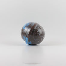 Load image into Gallery viewer, A mostly black colored, oval shaped bath bomb with splashes of blue on a white background
