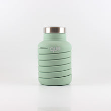 Load image into Gallery viewer, A photo of the compressed version of an expandable dusty sage water bottle with a metal cap, that says &quot;que&quot; in white lowercase letters.