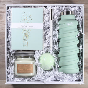 Overhead view of the "For Your Enjoy-Mint" gift box, which includes a light green water bottle, a light green bath bomb, a white pen with gold accents, a mason jar candle with green wax, and a light green journal that says "bucket list", all in a white box with white crinkle paper