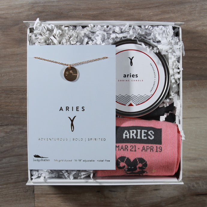 small aries gift box with an aries disc necklace, an aries candle, and aries socks. These gifts are in a white gift box with white crinkle paper.