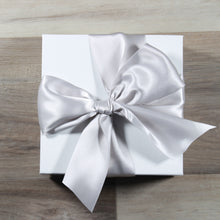 Load image into Gallery viewer, An overhead photo of a Doromania gift box - a white square gift box with a silver satin bow on a wood background.
