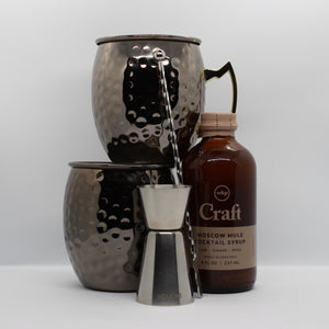 Contents of the "Mule it Over" gift box on a white background. 2 black hammered mugs, a silver double jigger, a silver swizzle stick, and a bottle of craft moscow mule cocktail syrup