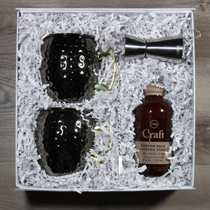 The "Mule it over" moscow mule gift box with two blakc hammered mugs, a silver double jigger, a silver swizzle stick, and a bottle of craft moscow mule cocktail syrup. Gift items are packaged in a white box with white crinkle paper.