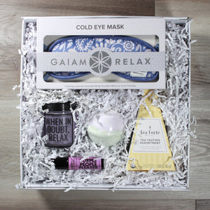 Overhead view of the "Major Chill Pill" gift box with a tea pyramid, a candle, an eye mask, an aromatherapy roller, and  a bath bomb, all in a white box with white crinkle paper