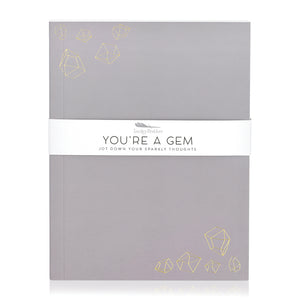 A purple-grey journal with gold embossed illustrations of crystal gemstones. A band around the journal says "You're a gem, jot down your sparkly thoughts."
