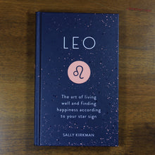 Load image into Gallery viewer, A dark blue book titled Leo: The art of living well and finding happiness according to your star sign. The book is by Sally Kirkman. The cover has the title, author, and the Leo zodiac symbol in a pink circle.