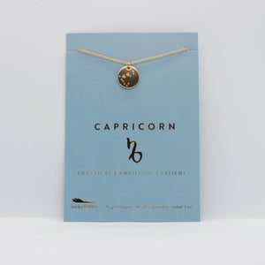 Capricorn Zodiac Necklace by Lucky Feather. A gold disc necklace with the Capricorn constellation etched in it, on a blue paper board.