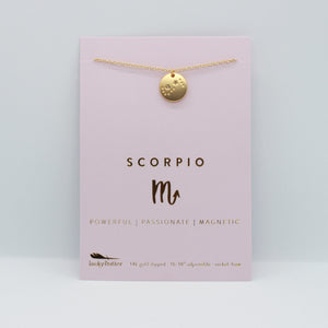 Scorpio zodiac necklace from Lucky Feather. A gold disc necklace with the scorpio constellation engraved on it. The necklace is packaged on a pink necklace display card.